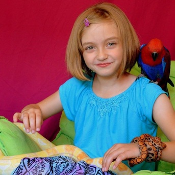 A child is sitting in a chair with a parrot on her shoulder, one snake wrapped around her wrist, and another snake in her lap.